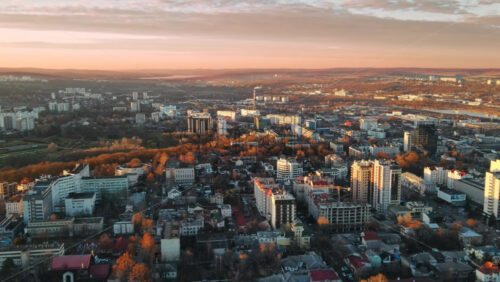 Video - Aerial drone view of Chisinau at sunset. Panorama view of multiple buildings, roads with moving cars, bare trees, park, lake and hills in the distance. Moldova