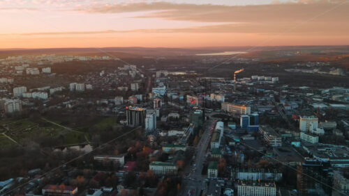 Video - Aerial drone view of Chisinau at sunset. Panorama view of multiple buildings, roads with moving cars, bare trees, park, lake and hills in the distance. Moldova