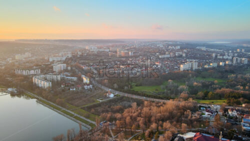 Video - Aerial drone view of Chisinau at sunset. Panorama view of multiple residential buildings, road with cars, bare trees, two parks and lake. Moldova