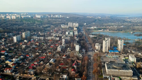 Video - Aerial drone view of Chisinau. Panorama view of multiple buildings, road with moving cars, bare trees, lake and hills in the distance. Good weather with fog in the air. Moldova