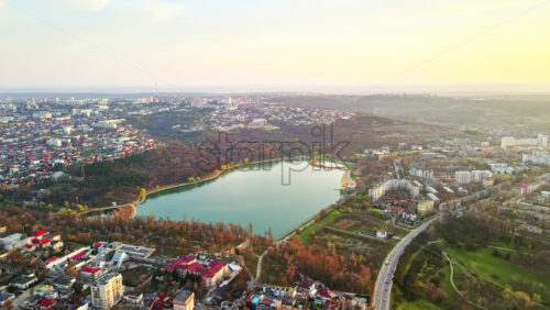 Video - Aerial drone view of Chisinau at sunset. Multiple buildings, trees, park with a lake, road with moving cars, setting sun. Moldova