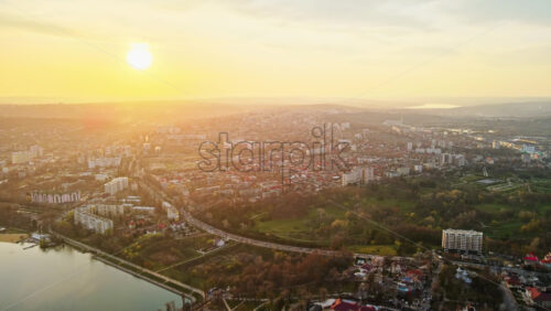 Video - Aerial drone view of Chisinau at sunset. Multiple buildings, trees, park with a lake, road with moving cars, setting sun. Moldova