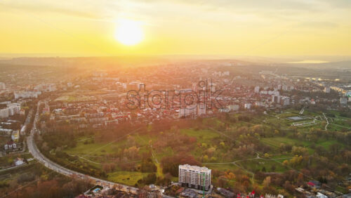Video - Aerial drone view of Chisinau at sunset. Multiple buildings, trees, park, road with moving cars, setting sun. Moldova