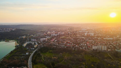 Video - Aerial drone view of Chisinau at sunset. Multiple buildings, trees, park with a lake, road with moving cars, setting sun. Moldova