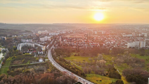 Video - Aerial drone view of Chisinau at sunset. Multiple buildings, trees, park with a lake, road with moving cars, setting sun. Moldova