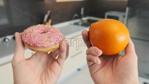 Video - Two male hands holding a donut and an orange. Chosing healthy food. Kitchen on the background