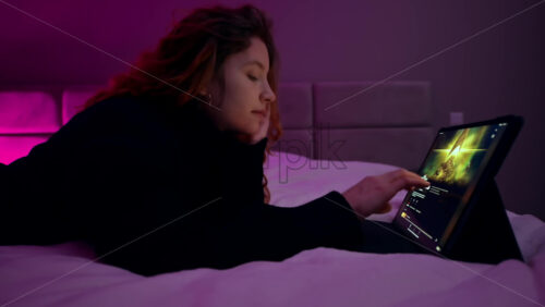 Video - CHISINAU, MOLDOVA - JANUARY 27, 2020: Young girl sitting in the bedroom with colorful light and browsing Netflix on tablet
