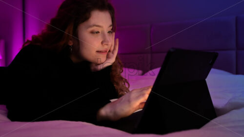 Video - Young ginger hair girl browsing her tablet at night in the bedroom. Colorful light on background
