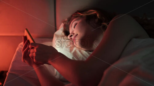 Video - Young ginger hair girl browsing her smartphone at night in the bed. Colorful light
