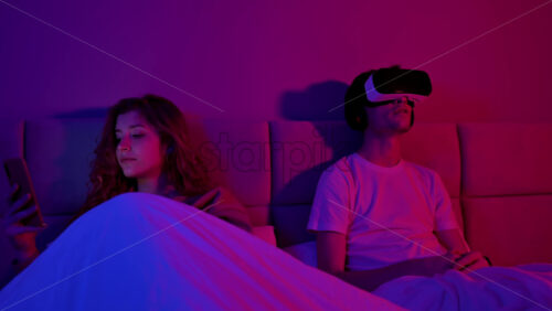 Video - Young couple in the bedroom at night before sleeping. Man using virtual reality headset, girl browsing smartphone. Colorful lights