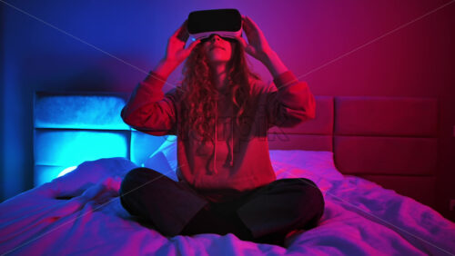 Video - Young girl sitting on the bed at night, using virtual reality headset. Neon colorful lights