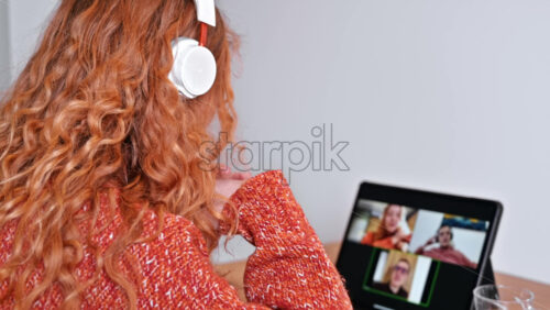 Video - Young ginger girl with headphones talking to her friends in video conference. Group of young people working from home