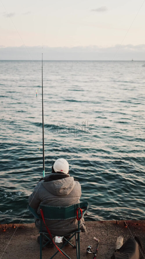 Video - Man in winter clothes on a chair fishing in the sea in the south of France. Vertical