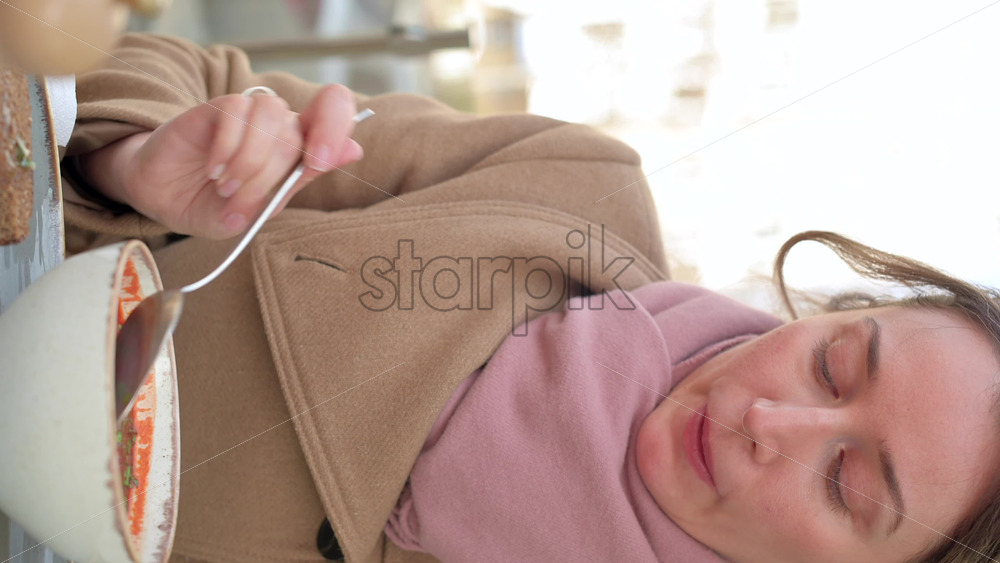 Video - Woman in brown coat eating borscht at a terrace. Vertical