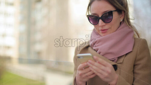 Video - Brunette woman wearing a brown coat scrolling on her phone at a terrace
