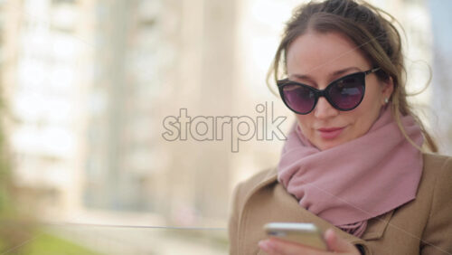 Video - Brunette woman wearing a brown coat scrolling on her phone at a terrace