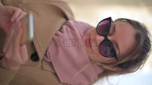 Video - Brunette woman wearing a brown coat scrolling on her phone at a terrace. Vertical