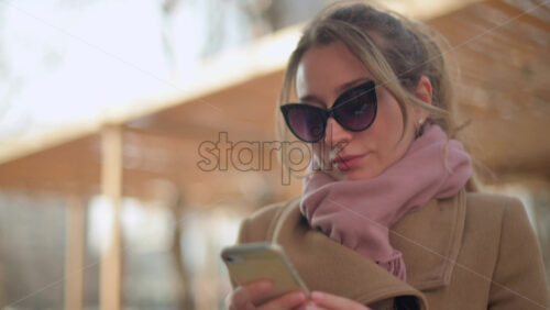 Video - Brunette woman wearing a brown coat scrolling on her phone at a terrace
