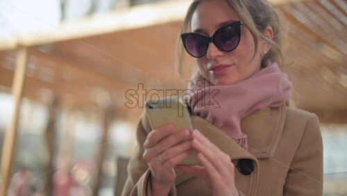 Video - Brunette woman wearing a brown coat scrolling on her phone at a terrace