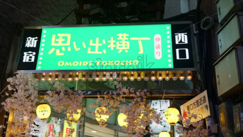 Video - Shinjuku, Japan - April 12, 2025: Lighted green Omoide Yokocho sign on the street with paper lanterns and cherry blossom flower decorations under it. Translation: " East exit"