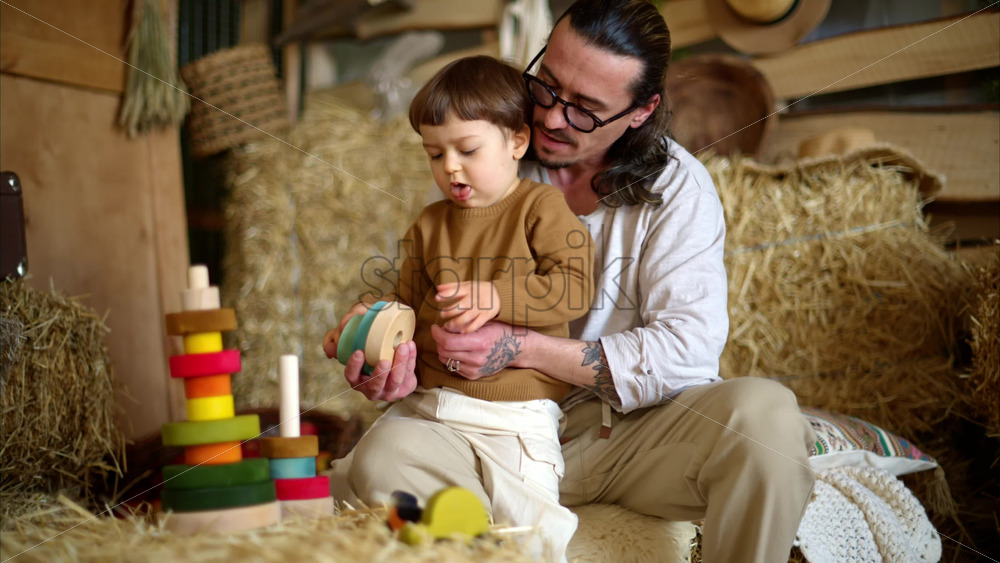 Video - Father playing with his son with colourful, ecological wooden toys in a barn, near square hay bales
