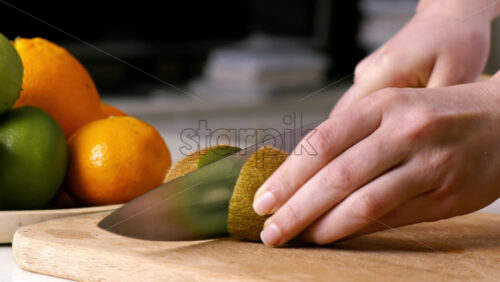 Video - Woman slicing kiwi in two parts on a wooden board. Fruits on the background. Slow motion