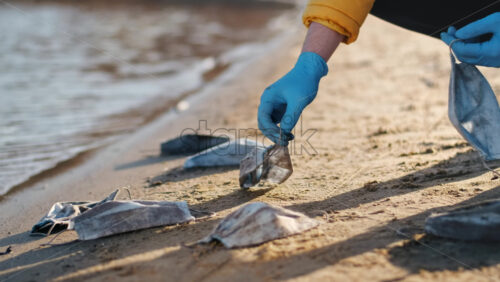 Video - Man in medical gloves picking up a bunch of dirty medical masks from the beach of a lake. Pollution idea