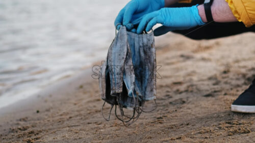 Video - Man in medical gloves picking up a bunch of dirty medical masks from the beach of a lake. Pollution idea