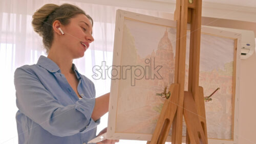 Video - Caucasian woman painting a picture with a palette in her hands and listening music with earphones indoors