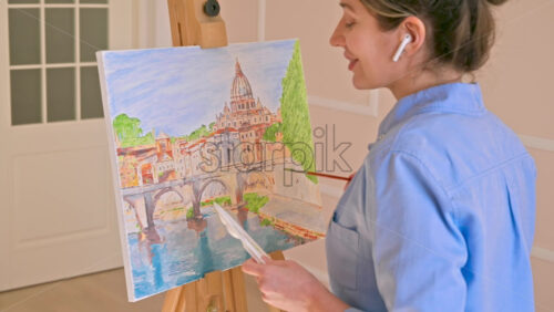 Video - Caucasian woman painting a picture with a palette in her hands and listening music with earphones indoors. Slow motion