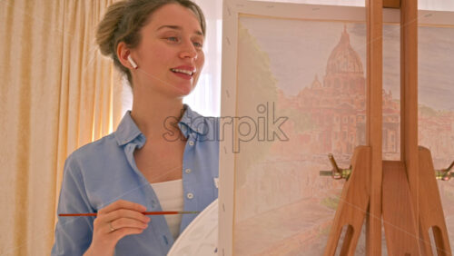 Video - Caucasian woman painting a picture with a palette in her hands and listening music with earphones indoors