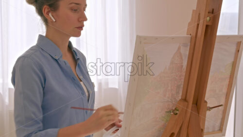 Video - Caucasian woman painting a picture with a palette in her hands and listening music with earphones indoors