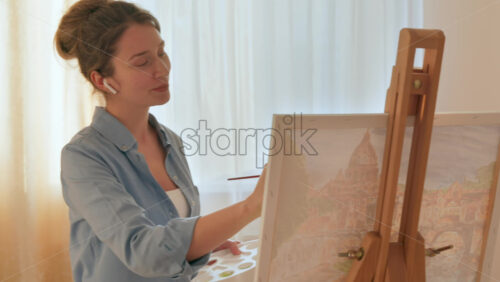 Video - Caucasian woman painting a picture with a palette in her hands and listening music with earphones indoors