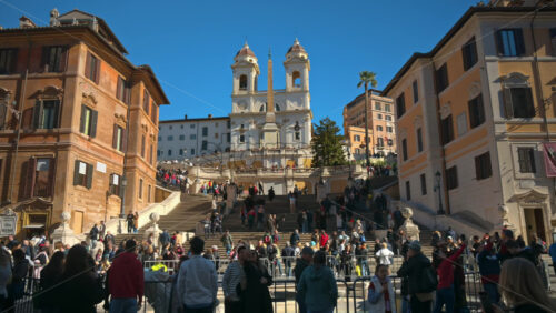 Video - Rome, Italy - February 14, 2024: Tourists walking on Spanish square near the baroque-style Fountain of the Boat in sunlight. Church of Santissima Trinità dei Monti in background