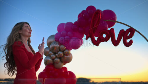 Video - Beautiful young woman in red dress taking photos of the sunset near the red and pink balloon arch spelling out the word "love"