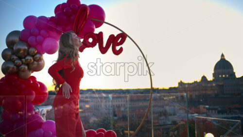 Video - Beautiful young woman in red dress dancing at sunset near the red and pink balloon arch spelling out the word "love". Vatican city in background. Rome, Italy