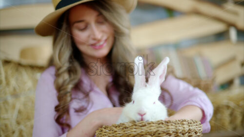 Video - Woman petting a white bunny in the barn near square hay bales, in daylight