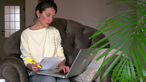 Video - Caucasian woman sitting on a chair, working on her laptop and making notes in notepad