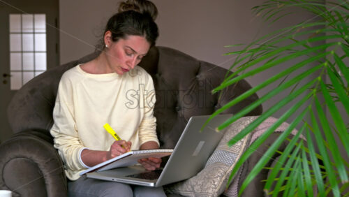 Video - Caucasian woman sitting on a chair, working on her laptop and making notes in notepad