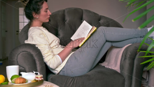 Video - Caucasian woman sitting on a chair and reading a book. Table with tea and fruits nearby