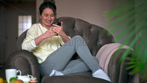Video - Caucasian woman sitting on a chair is on her smartphone and smiling. Table with tea and fruits nearby