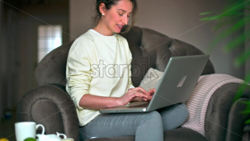 Video - Happy caucasian woman sitting on a chair and working on her laptop. Table with tea and fruits nearby