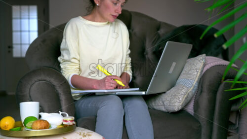 Video - Caucasian woman sitting on a chair and is at online meeting using laptop, talking and making notes in notepad. Cat, table with tea and fruits nearby