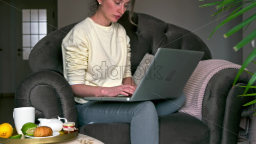Video - Caucasian woman sitting on a chair and working on her laptop. Table with tea and fruits nearby