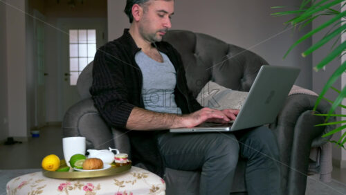 Video - Caucasian man sitting on a chair and working on his laptop. Table with tea and fruits nearby
