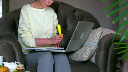 Video - Caucasian woman sitting on a chair, working on her laptop and making notes in notepad. Table with tea and fruits nearby