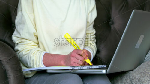 Video - Caucasian woman sitting on a chair, working on her laptop and making notes in notepad