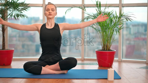 Video - Young woman in yoga lotus position meditating indoors in apartment with city on background