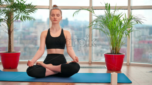 Video - Young woman in yoga lotus position meditating indoors in apartment with city on background