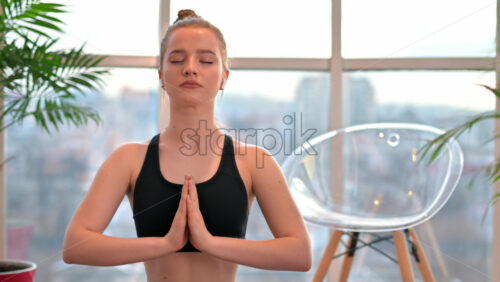 Video - Young woman in yoga prayer pose meditating indoors in apartment. City on background.